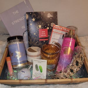May include: A gift basket filled with self-care items. Includes a book titled "The Strength In Our Souls", a journal, a candle, a body spray, a lip balm, a jar of bath salts, a small jar of cream, a decorative jar, a small crown, and a card.