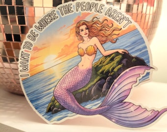 May include: A sticker featuring a mermaid illustration. The mermaid has long red hair, a seashell top, and a purple tail. The background shows a sunset over the ocean with the text "I WANT TO BE WHERE THE PEOPLE AREN'T".