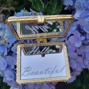 May include: A gold-colored, rectangular compact mirror with an open lid. The mirror is engraved with the words "Hello Beautiful" in cursive script. The compact is set against a backdrop of blue hydrangea flowers.