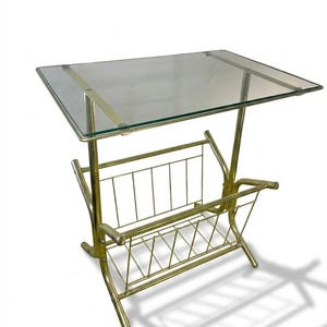 May include: A vintage brass-toned side table with a clear glass top and a magazine rack. The table has a rectangular glass surface and a lower rack with a geometric design. The legs are curved and the overall design is mid-century modern.