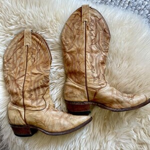 May include: A pair of tan cowboy boots with intricate stitched designs. The boots have pointed toes, stacked heels, and pull straps. The leather shows wear and a vintage aesthetic, suggesting they are well-loved and broken in.