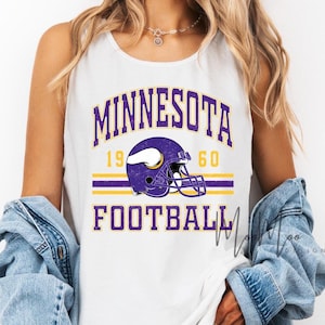 May include: White tank top featuring the text "MINNESOTA FOOTBALL" in purple and gold. A purple football helmet graphic is centered above the word "FOOTBALL". The year "1960" is above the helmet. The tank top is sleeveless.