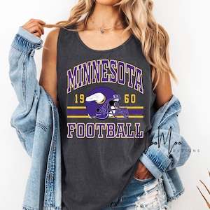 May include: Dark gray tank top featuring the text "MINNESOTA FOOTBALL" in purple and white, with a purple football helmet graphic. The year "1960" is also displayed. The design includes yellow stripes.