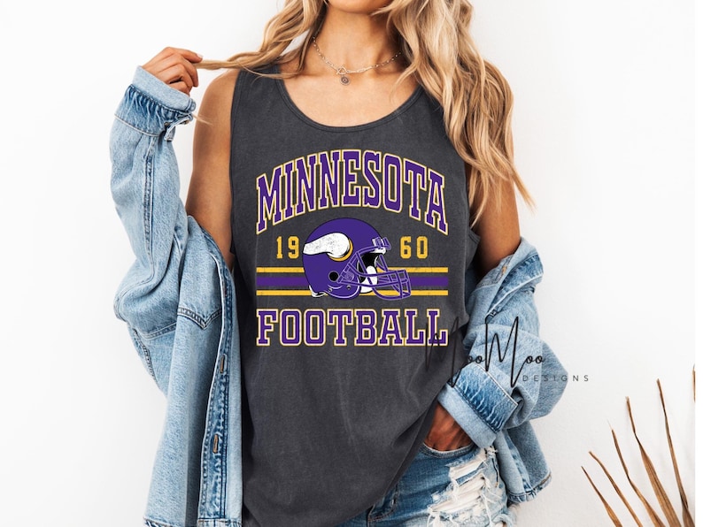 May include: Dark gray tank top featuring the text "MINNESOTA FOOTBALL" in purple and white, with a purple football helmet graphic. The year "1960" is also displayed. The design includes yellow stripes.