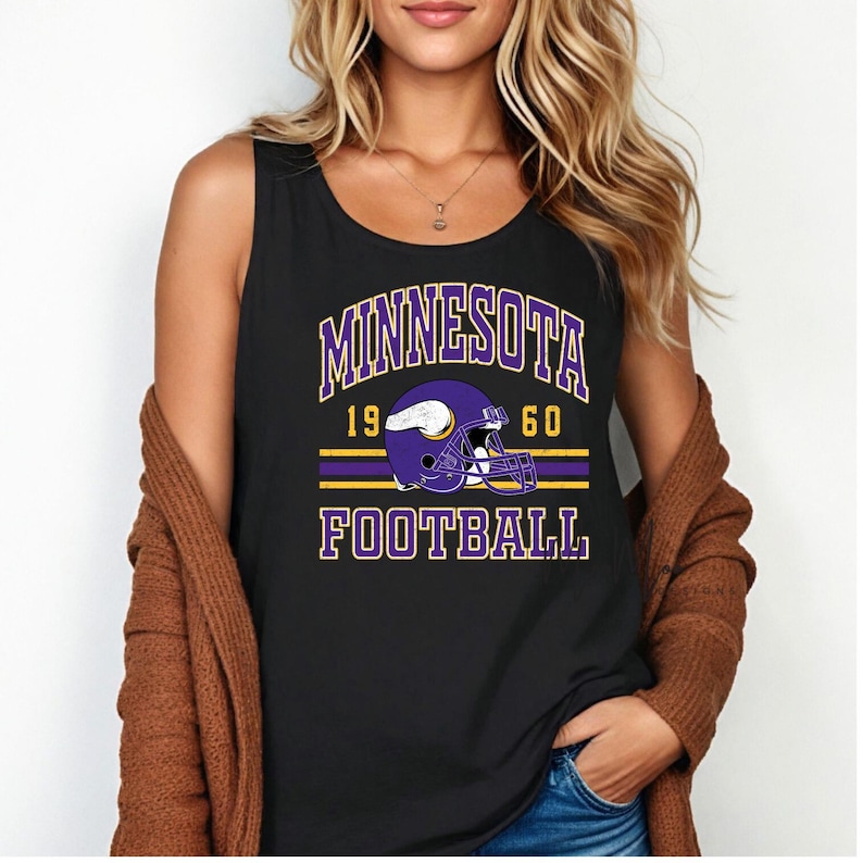 May include: Black tank top featuring the text "MINNESOTA FOOTBALL" in purple and gold. The design includes a purple football helmet with a white outline. The year "1960" is printed above the helmet.
