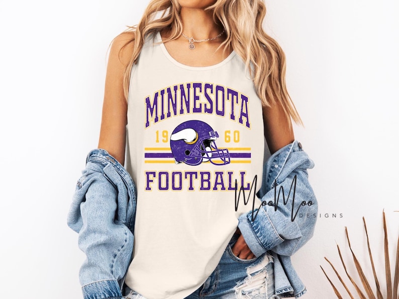 May include: Cream-colored tank top featuring the words "MINNESOTA FOOTBALL 1960" in purple and gold. A purple football helmet graphic is centered on the shirt. The tank top is sleeveless.