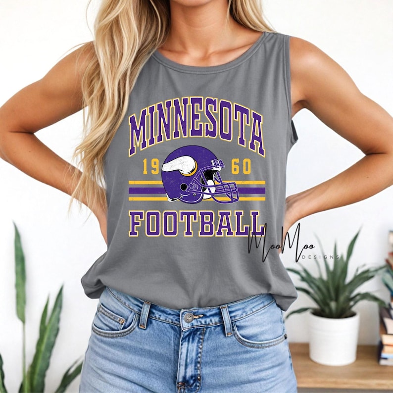 May include: Gray tank top with the words "MINNESOTA FOOTBALL" in purple and gold. The design includes a purple football helmet and the numbers "1960". The shirt is sleeveless and made of a soft material.