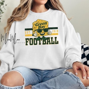 May include: White sweatshirt featuring a cartoon cheese wedge wearing sunglasses and giving a thumbs up. The design includes the words "Green Bay Football" and "Est. 1919" in green and yellow.