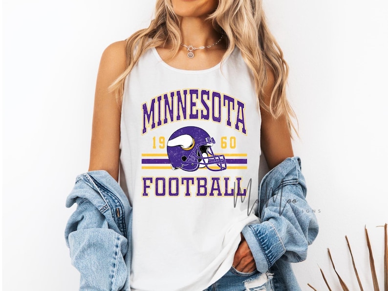 May include: White tank top featuring the text "MINNESOTA FOOTBALL" in purple and gold. A purple football helmet graphic is centered above the word "FOOTBALL". The year "1960" is above the helmet. The tank top is sleeveless.