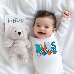 May include: A smiling baby wearing a white onesie with the word "BUBS" in teal, orange, and blue letters. The onesie has a checkered pattern and smiley faces. A plush teddy bear and a card that says "Hello" are also in the image.