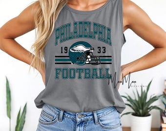 May include: Grey vest top with the words "PHILADELPHIA FOOTBALL" in teal, and a football helmet graphic. The shirt also has the numbers "19" and "33". A casual, sporty design.