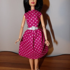 May include: A pink and white polka dot dress with a white belt and a white bow. The doll is wearing white shoes.