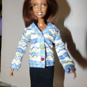 May include: A doll wearing a blue, yellow, and white patterned sweater with a black skirt. The doll has brown hair and brown eyes.