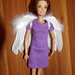 May include: A doll with brown hair and a purple dress is wearing white angel wings. The doll has blue eyes and a light pink blush on her cheeks.