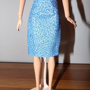 May include: A blue pencil skirt with a white floral pattern. The skirt is knee-length and has a fitted silhouette. The doll is standing on a white stand.