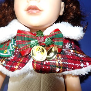 May include: A Christmas-themed doll cape with a red and green plaid pattern, white faux fur trim, and a decorative bow. The cape features a Christmas tree patch and two golden bells.