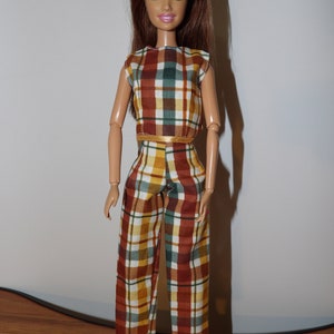 May include: A doll wearing a plaid top and pants. The top and pants are brown, yellow, green, and white. The doll has long brown hair and blue eyes.
