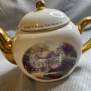 May include: White ceramic teapot with gold accents, including the handle, spout, and lid knob. The teapot features a painted oval scene of a house and the text "Home Is Where The Heart Is" on the lid.