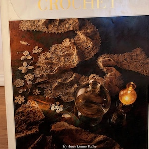 May include: A book titled "A Living Mystery The International Art & History of Crochet" with gold lettering. The cover features a dark background with crochet lace and a glass bottle. The author's name, Annie Louise Potter, is at the bottom.