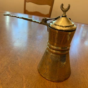 May include: A vintage brass coffee pot with an etched design. The pot has a long handle and a lid with a crescent-shaped finial. The pot is sitting on a wooden surface.