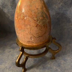 May include: A large, polished, egg-shaped stone with a mottled peach and tan pattern. The stone sits on a small, ornate, brass-colored metal stand with curved legs. The egg has visible cracks.