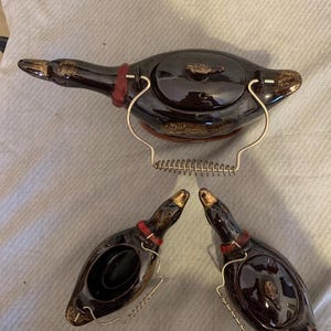 May include: A dark brown ceramic duck-shaped teapot with a matching lid and two duck-shaped salt and pepper shakers. The teapot and shakers have gold accents on the beak and wings, and a red band around the neck. The teapot has a metal handle.