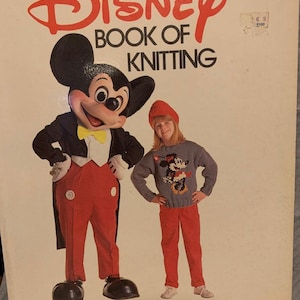 May include: The Disney Book of Knitting cover featuring Mickey Mouse and a girl in a knitted Mickey Mouse sweater. The book's title is in red, with the authors' names at the bottom. The girl is wearing a red hat and pants.