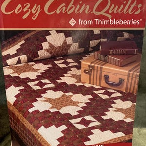 May include: A book titled "Cozy Cabin Quilts" by Lynette Jensen, featuring a quilt with a star pattern in shades of brown, white, and tan. The book cover is red with gold lettering. The image also shows a stack of books and a small suitcase.