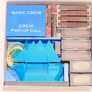 Peut inclure: Un organisateur de jeu de société rempli de composants de jeu. La boîte contient des inserts en mousse bleue avec le texte "BASIC CREW" et "PORT OF CALL". Il y a aussi des jeux de cartes, des jetons et une pièce de jeu en forme de bateau bleu. La boîte est blanche.