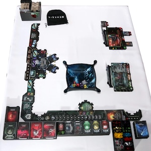 May include: A board game setup with various components on a white surface. Includes game boards, cards, tokens, and a black bag with "NEMESIS" printed on it. The game pieces are in shades of black, green, red, and blue.