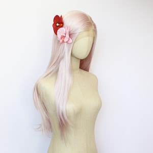 May include: A mannequin head with long, pale pink hair decorated with two fabric flower hair clips. One clip is red with a pearl centre, and the other is pink. The mannequin is against a white backdrop.