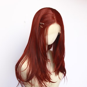 May include: A long, auburn wig with a center part, styled on a mannequin head. The wig features a natural-looking hairline and long, layered strands. Two small gold hair clips are placed near the part. The background is plain white.