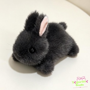 May include: A small plush rabbit toy covered in dark gray faux fur with a soft texture. The rabbit has two upright ears, one of which shows a light pink inner lining, and a small round tail. It features black plastic eyes and thin whiskers.