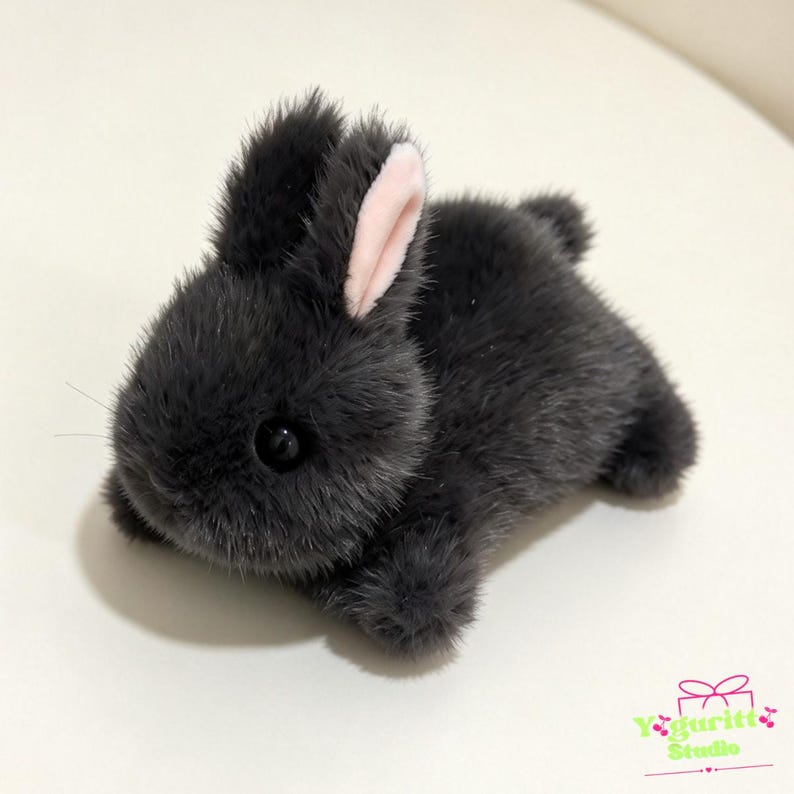 May include: A small plush rabbit toy covered in dark gray faux fur with a soft texture. The rabbit has two upright ears, one of which shows a light pink inner lining, and a small round tail. It features black plastic eyes and thin whiskers.