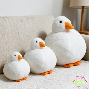 May include: Three round white plush duck toys of varying sizes, each featuring a soft white body, a bright orange beak, and small orange feet. The ducks have black bead eyes and a smooth, rounded shape, arranged in a row from smallest to largest.