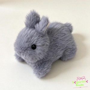May include: A small, plush rabbit figurine covered in soft, light gray faux fur. The rabbit has two upright ears, a rounded body, a short tail, and two shiny black bead eyes. In the bottom right corner, there is a logo with the text Yguritt Studio.