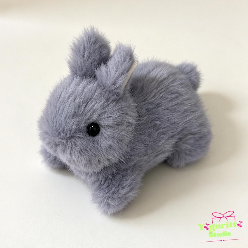 May include: A small, plush rabbit figurine covered in soft, light gray faux fur. The rabbit has two upright ears, a rounded body, a short tail, and two shiny black bead eyes. In the bottom right corner, there is a logo with the text Yguritt Studio.