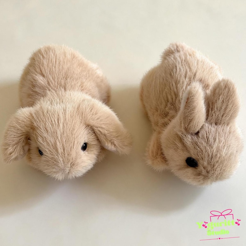 May include: Two plush stuffed animals shaped like rabbits with soft, light beige fur. One rabbit features long, floppy ears hanging down, while the other rabbit has upright, pointed ears. Both have small, dark, round eyes and a rounded body shape.