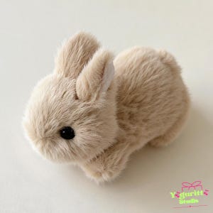 May include: A small, plush rabbit figurine covered in soft, light beige faux fur. The rabbit has two upright ears with white inner lining, a rounded body, and a single visible black eye. The text Yaguritt Studio is printed in the bottom corner.