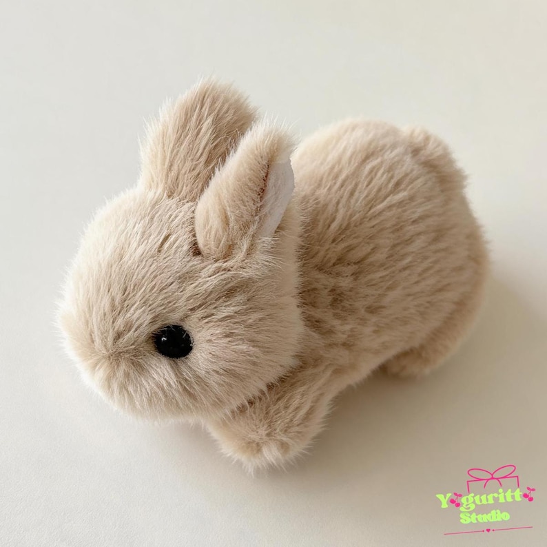 May include: A small, plush rabbit figurine covered in soft, light beige faux fur. The rabbit has two upright ears with white inner lining, a rounded body, and a single visible black eye. The text Yaguritt Studio is printed in the bottom corner.