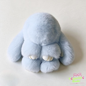 Puede incluir: Un juguete de peluche de conejo azul claro con pelaje largo y suave y dos patas blancas con detalles cosidos en color dorado. El animal de peluche tiene una cabeza redondeada y orejas largas y caídas, con el texto Yoguritt Studio visible.