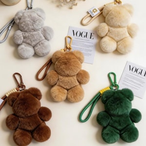 Op de afbeelding: Een verzameling pluche teddybeer sleutelhangers in verschillende kleuren: grijs, beige, bruin, donkerbruin en groen. Elke beer heeft een goudkleurige sluiting en een bijpassende polsband. Het woord "VOGUE" is zichtbaar op een tijdschriftenpagina.