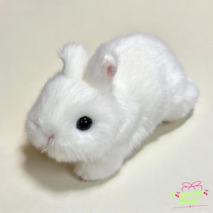 May include: A small, plush toy rabbit covered in soft, white faux fur. The rabbit has two upright ears with a hint of pink on the inner side, a small pink nose, and large, round, black plastic eyes. The figure is positioned in a resting pose.