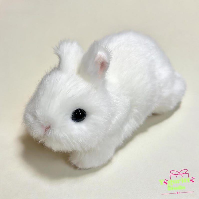 May include: A small, plush toy rabbit covered in soft, white faux fur. The rabbit has two upright ears with a hint of pink on the inner side, a small pink nose, and large, round, black plastic eyes. The figure is positioned in a resting pose.