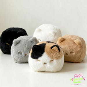 Könnte beinhalten: Eine Sammlung von fünf runden, katzenförmigen Plüschkissen aus weichem Kunstfell. Die Katzen sind schwarz, weiß, grau, hellbraun und eine Glückskatze mit schwarzen, orangefarbenen und weißen Flecken. Jedes Plüschtier hat gestickte Augen und eine Nase.