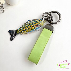 May include: A keychain featuring a segmented, articulated fish charm with yellow, blue, and black stripes, attached to a lime green rectangular strap and a dark metallic circular ring. The fish has a dark tail and small metal loops on its underside.