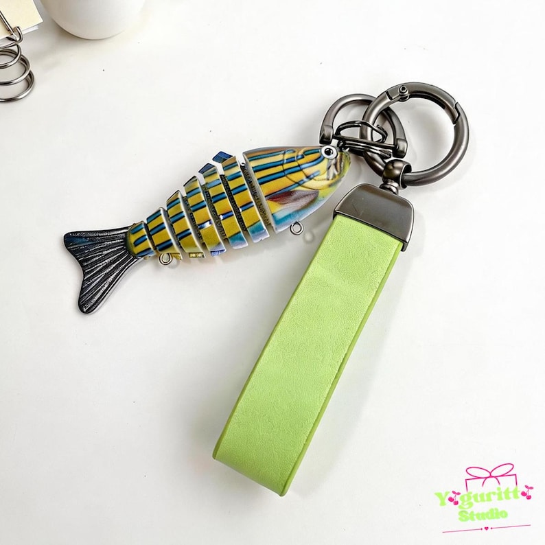 May include: A keychain featuring a segmented, articulated fish charm with yellow, blue, and black stripes, attached to a lime green rectangular strap and a dark metallic circular ring. The fish has a dark tail and small metal loops on its underside.