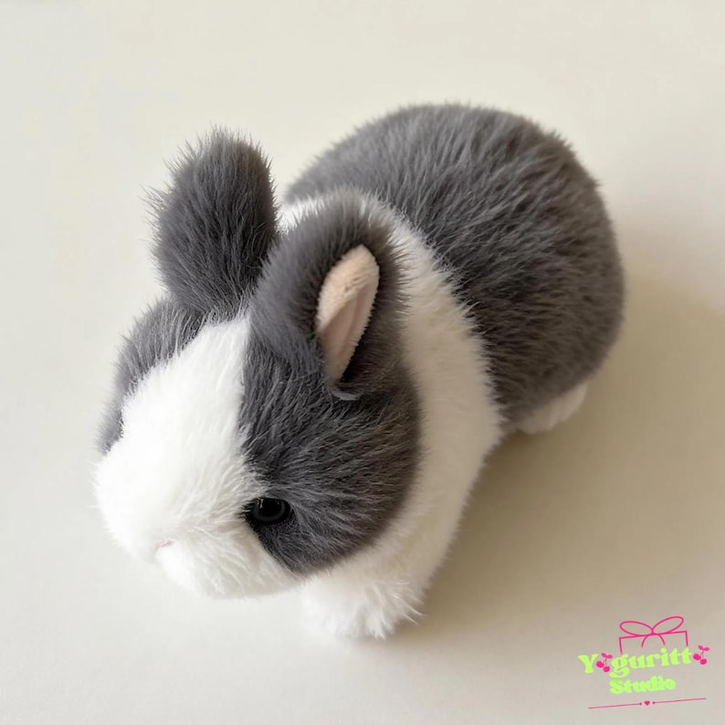 May include: A plush toy rabbit featuring soft, faux fur in a two-tone pattern of dark gray and white. The rabbit has upright ears with light pink inner lining, dark round eyes, and a small nose. The text Yoguritt Studio is visible in the corner.