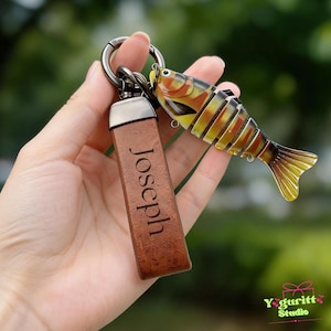 May include: A keychain featuring a brown rectangular leather strap engraved with the name Joseph and a multi-segmented, articulated fish lure with yellow, orange, and black stripes, all attached to a dark metal circular ring.
