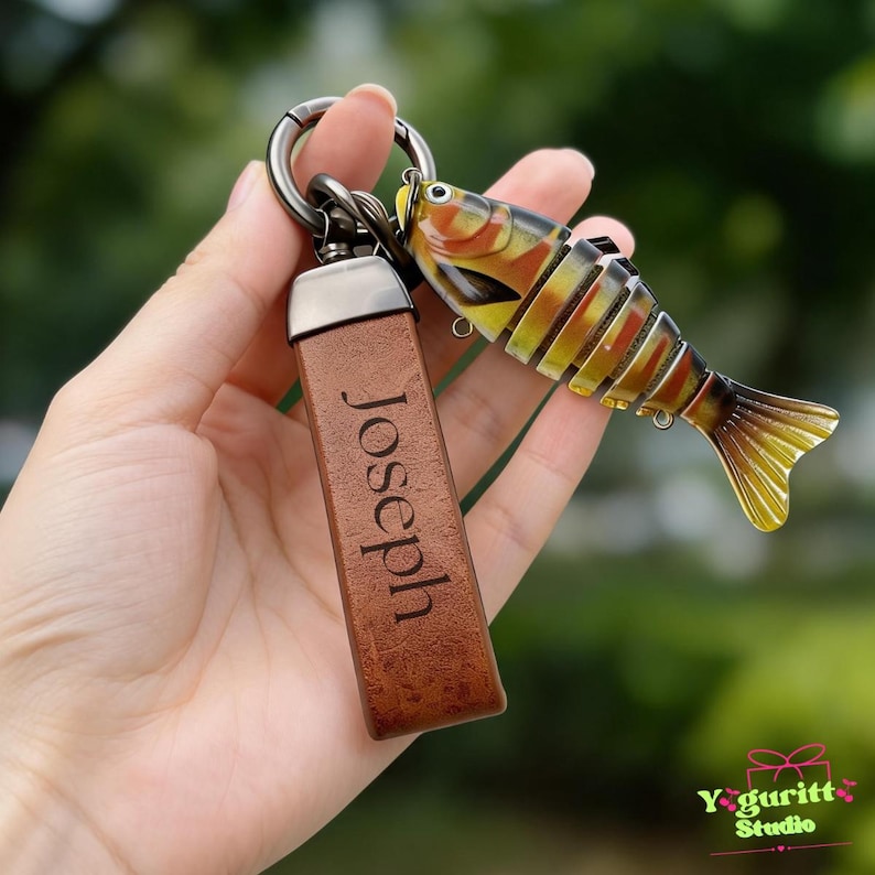 May include: A keychain featuring a brown rectangular leather strap engraved with the name Joseph and a multi-segmented, articulated fish lure with yellow, orange, and black stripes, all attached to a dark metal circular ring.